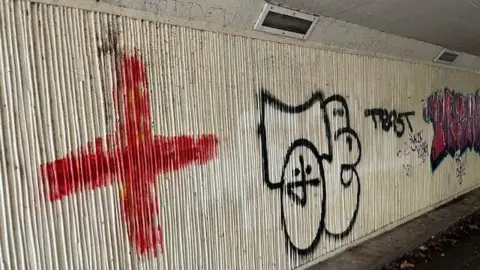 John Edwards Photo of the graffiti in the underpass, which has rough cream walls. There are three pieces of graffiti visible, from left to right: a large red cross, a stylised 'tag' reading 'JE', and a large purple piece of text that is not legible. Around the larger pieces are various scribbles in black ink. 