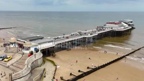An aerial view of Cromer pier stretching out to sea. People are milling about on the pier and the beach below, and the sea is calm. 