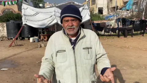 Abu Ahmed stands in a cream-coloured jacket zipped partly open at the front and a dark blue woollen hat, his hands held out, in front of a makeshift tent, buildings and clothes on a line.