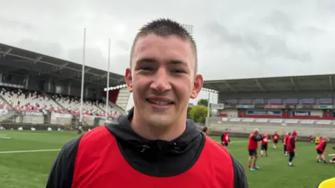 A young man standing in front of a rugby training session in a stadium wearing a red bib, black hoody and smiling. He has brown spiky hair and his team mates behind him are wearing red bibs.