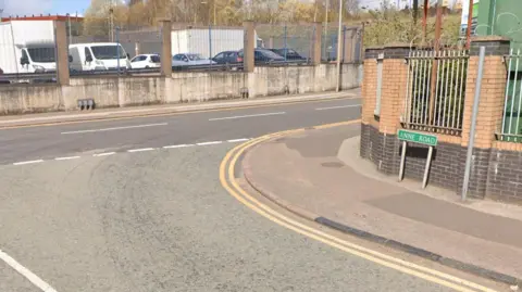 Google A Google street view image of the T-junction at Anne Road and Booth Street. A green sign saying Anne Road is on the right in front of a fence with brick pillars. A car park is visible behind a fence in the background.