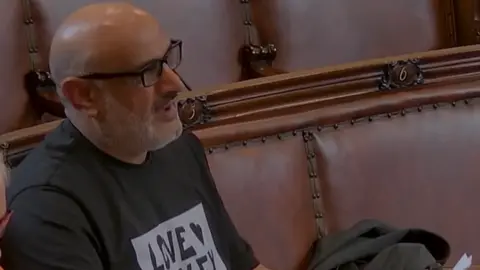 Wakefield Council Ayub seated on one of the benches. He has a bald head, wears rectangular black-framed glasses, and has closely trimmed facial hair. He is dressed casually in a dark T-shirt with partially visible white text that reads “LOVE …” across the chest. He is turned slightly to the side, appearing to be looking toward something or someone out of frame.