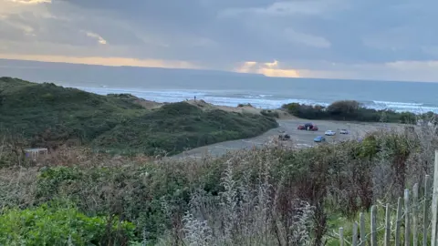 Saunton Sands is seen in the background, with waves seen beneath an area of sunlight emerging from the clouds. In the foreground is a car park and shrubbery, with dunes to the left. 