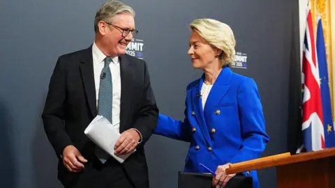 Carl Court/Getty Images Prime Minister Keir Starmer, wearing a suit, tie, dress shirt and glasses, smiles as he looks at European Commission, Ursula von der Leyen, also wearing a suit. Her lips are upturned and her hand is on his back. They are walking away from a podium at a press conference, holding notes. To the right in the background are a British and European Union flag.