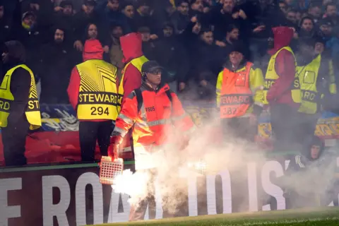 EPA/Shutterstock A man in an orange jacket stood with two lit flares in a red steel cage. He is stood on the side of a football pitch with fans and stewards behind him.
