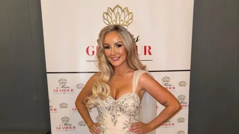 Emily Kathleen The image shows a woman in a gold sparkly dress standing in front of a cream backdrop featuring a crown and the words "Miss Glamour UK".