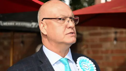 A bald man with glasses, wearing a black jacket, light blue tie and white shirt, looks to his left.