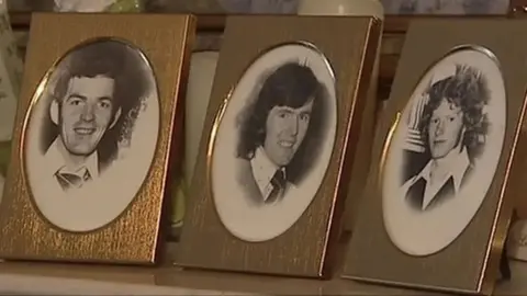 Three individual framed pictures of brothers Anthony, John Martin and Brian Reavey sit on top of  a table. The pictures are black and white. John Martin has short dark hair and is smiling, Brian has long dark hair and is smiling while Anthony has curly, long lighter hair 