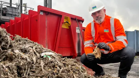 Jonah Fisher/BBC Un empleado de Severn Trent sostiene una toallita húmeda sucia frente a una pila de toallitas húmedas. 