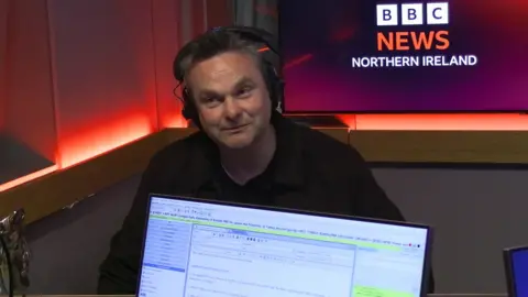 A man with headphones on in a radio studio. In the foreground, in front of him, we can see the front of a computer screen used by the radio programme presenter. In the background is a screen showing the BBC News Northern Ireland logo