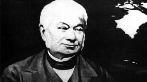 University of Lancashire A black and white portrait of Livesey. He has white hair and white sideburns, He wears a black jacket, a white high-collared shirt