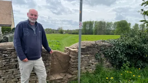 LDRS A man standing next to a stone wall. He's wearing a quarter zip blue jumper with white trousers and leaning his arm on the corner of the wall. There's a stone stile on the right-hand side of him next to where he's leaning, with the path stretching into a grass meadow. There are trees in the background and an overcast sky. To the right of the image, the stone wall starts to be enveloped in ivy until you can't see it at all towards the end of the picture. In front of the stone wall on the ground is a patch of grass and dandelions. There's a lamp post as well just in front of the wall. There's a house in the background on the left-hand side of the man