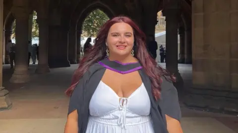 Maddy Dhesi A woman at graduation. She looks at the camera and smiles. She has a white dress and a black and purple graduation gown.