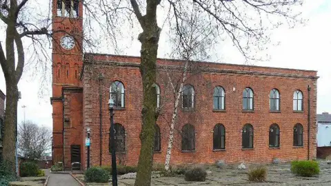 A two-storey redbrick building with a clocktower, called the St Thomas Centre, in Ardwick Green. The site can be seen from a paved courtyard with several trees in the foreground on an overcast day. 