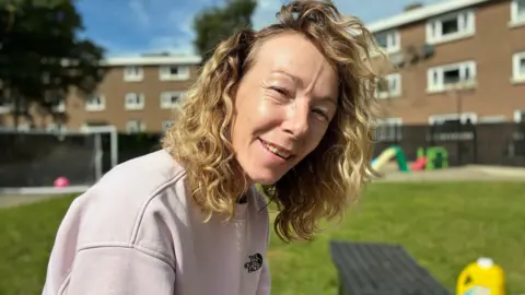 A woman with curly blonde hair smiles outdoors on a sunny day. She’s wearing a pale pink sweatshirt with a visible “The North Face” logo. Behind her, a grassy area features a football goal, colourful playground equipment, and residential buildings with white-framed windows. The bright light and relaxed pose create a friendly, community atmosphere.