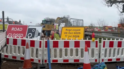 BBC The picture shows road closure red and white stripped fences behind two orange traffic cones. In the background are large metal construction fences and a yellow JCB digger.