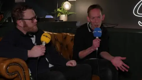 Huw Stephens and Steve Lamacq sit on a sofa at the venue holding 6 Music microphones.