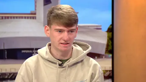 BBC Alfie, wearing a light grey hoodie, speaks in a BBC studio about his late father.