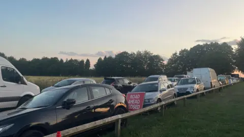 Residents Cars parked close together, on grass verges either side of a small country road.