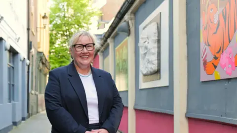 LDRS Head-and-shoulder view of Karen Kilgour, a smiling woman with tied black blonde hair, wearing spectacles, and a dark blue jacket over a white top. She is standing in what looks to be a lane or an alley and the building to her left is decorated with colourful artworks.