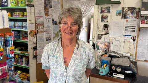 BBC A lady with grey short hair  standing in a shop. there are toothbrushes, petfood and a till in the background.