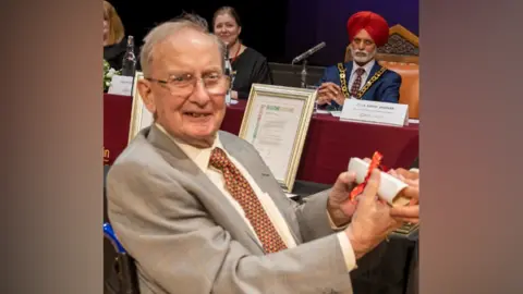 Telford & Wrekin Council Austin appears to be sitting in a council meeting and is turning around to the camera. He has a grey suit on and white shirt