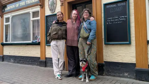 Dotty McLeod/BBC Three people are standing next to each other in front of the doorway of a pub. Jenni Skinner is on the left and is wearing a black T-shirt, khaki jacket and beige trousers. Amber Wenman is in the middle and has long pink hair and is wearing a red patterned T-shirt and black trousers. Niamh Sweeney is on the right and is wearing a denim jacket, grey top and green patterned trousers. There is a small beige and black dog. 