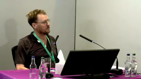 Stuart Ayris looks towards Baroness Lampard. He is wearing a black polo shirt and is sitting in front of a purple table with a laptop in front of him. He is wearing a green lanyard and has bottles of water in front of him. He is sitting in the inquiry room, with a microphone in front of him at Arundel House in central London