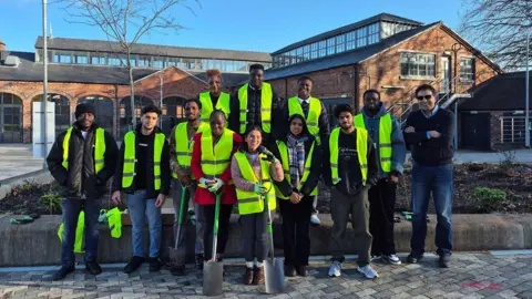 University of Wolverhampton Twelve people, most of them in hi-vis jackets, are together standing up facing the camera. Two of them at the front are holding spades and buildings are behind them.