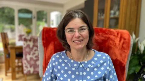 Seckin McGuirk at home in Rugby, Warwickshire, sitting in an armchair in a living room, wearing a blue dress with white spots