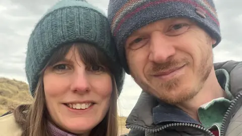 Annette Illing is standing next to her husband Mark on a beach on a cloud day. They are both wearing coats and knitted hats.