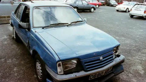 Pacemaker An archived photo of a blue car which has been damaged. A camera operator is filming the car.