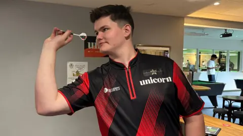 A young man with dark hair throwing a dart.