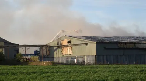Shot of a large green and grey-coloured industrial shed with plumes of smoke seen rising above it in the background. A fire service aerial ladder is visible in the photograph.