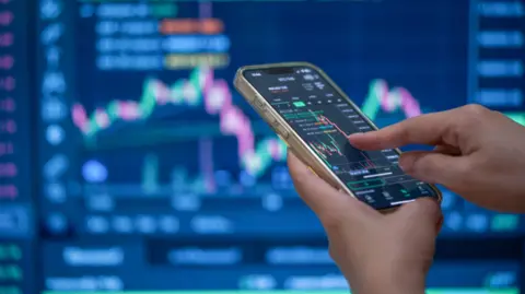 A trader's hands using their phone to make a trade in front of a trading screen in soft focus