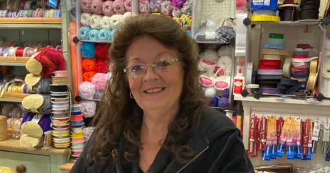 Local Democracy Reporting Service A woman with long dark hair and wearing glasses is smiling at the camera. There are shelves behind her from left to right and shelves directly behind her head on the photo include wool.