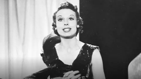 A black and white image from 1936 of a TV broadcast of a lady in evening dress singing. 