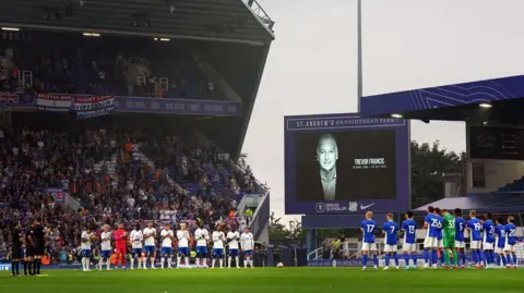 Jacob King/PA Wire Two football teams stand on a pitch, with fans on seats in the background. The team on the left is wearing white, the team on the left is wearing blue. There is a large TV screen between the two teams with a black and white photo of a man and the words "Trevor Francis". 