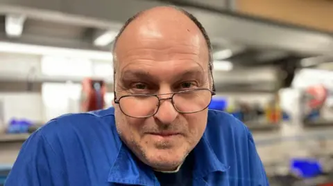 Helen Mulroy/BBC A man looking directly at the camera. He is wearing glasses and blue overalls.