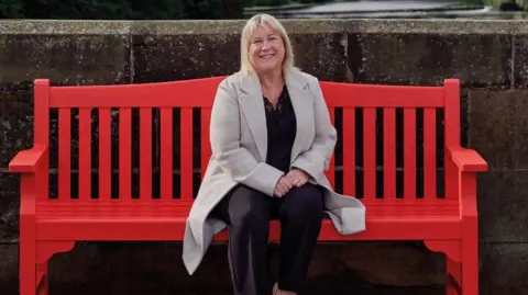 Louise McGill sitting on a large red bench. She has shoulder-length blonde hair, is smiling and wearing a light grey long coat over a dark top and trousers. Behind her is a high stone wall.