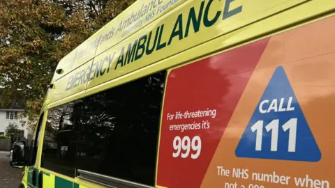 West Midlands Ambulance Service An ambulance with yellow high visibility livery, seen from the side, with trees and buildings in the distance in front of the vehicle.