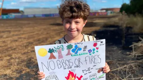 A young boy with brown curly short hair is smilingn and looking at the camera. He is holding a hand-made sign which says "no bbq's or fires in our parks" with other words and brightly coloured drawings, like trees and butterflies. Behind him is dry brown grass land. 