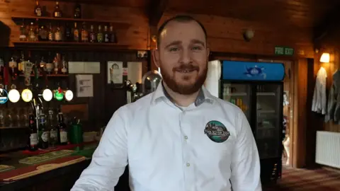 A man with short dark ginger hair and a short beard smiles at the camera while standing in a bar. He wears a white shirt 