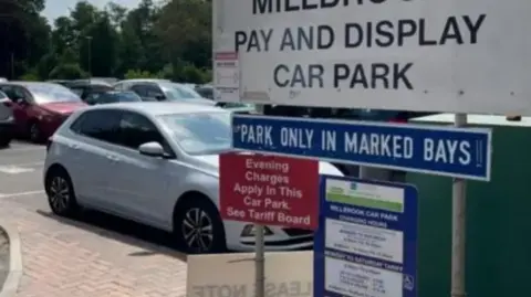 Surrey County Council A car park with signage advising of parking costs and to 'park in marked bays'. A white car is the most visible but there are other rows of cars behind. 