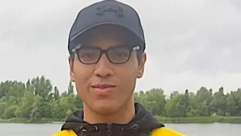 Facebook Nicholas Prosper dressed in glasses, a black baseball hat and yellow and black hoodie while standing in front of a lake.