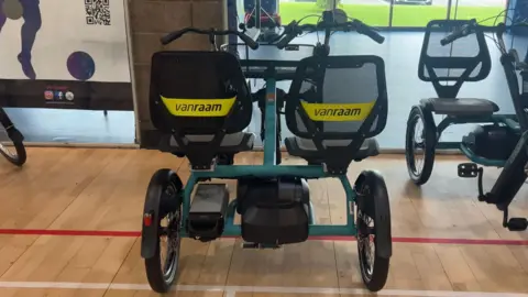 A close up of a side by side tandem tricycle made by Van Raam. There are two seats at the back of the bike with two wheels underneath. There are two handle bars with a basket in between them and one wheel underneath them. Below the double seats you can see the battery for the power assist.