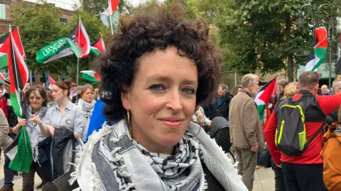 Orla Wallace smiling at the camera during the Belfast protest.  She has dark, curly hair and is weaing gold hooped earrings and a black and white scarf.  People behind her are carrying Palestinian flags. 