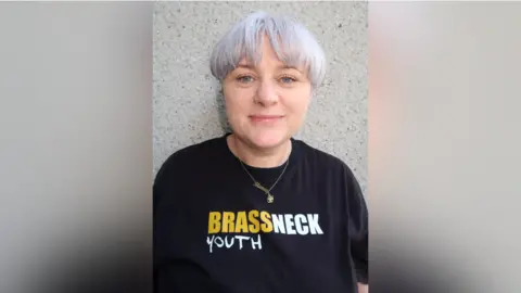 Brassneck Youth Theatre Alison is wearing a black t-shirt with a yellow and white logo saying Brassneck Youth Theatre. She has grey/purple short hair and is looking at the camera. 