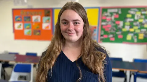 Katja has long brown hair and is wearing a blue jumper. She is standing in a classroom at her school.