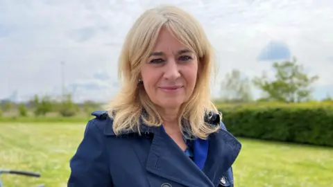 Christine Middlemiss is wearing a blue coat and is standing in a green field on a sunny day.